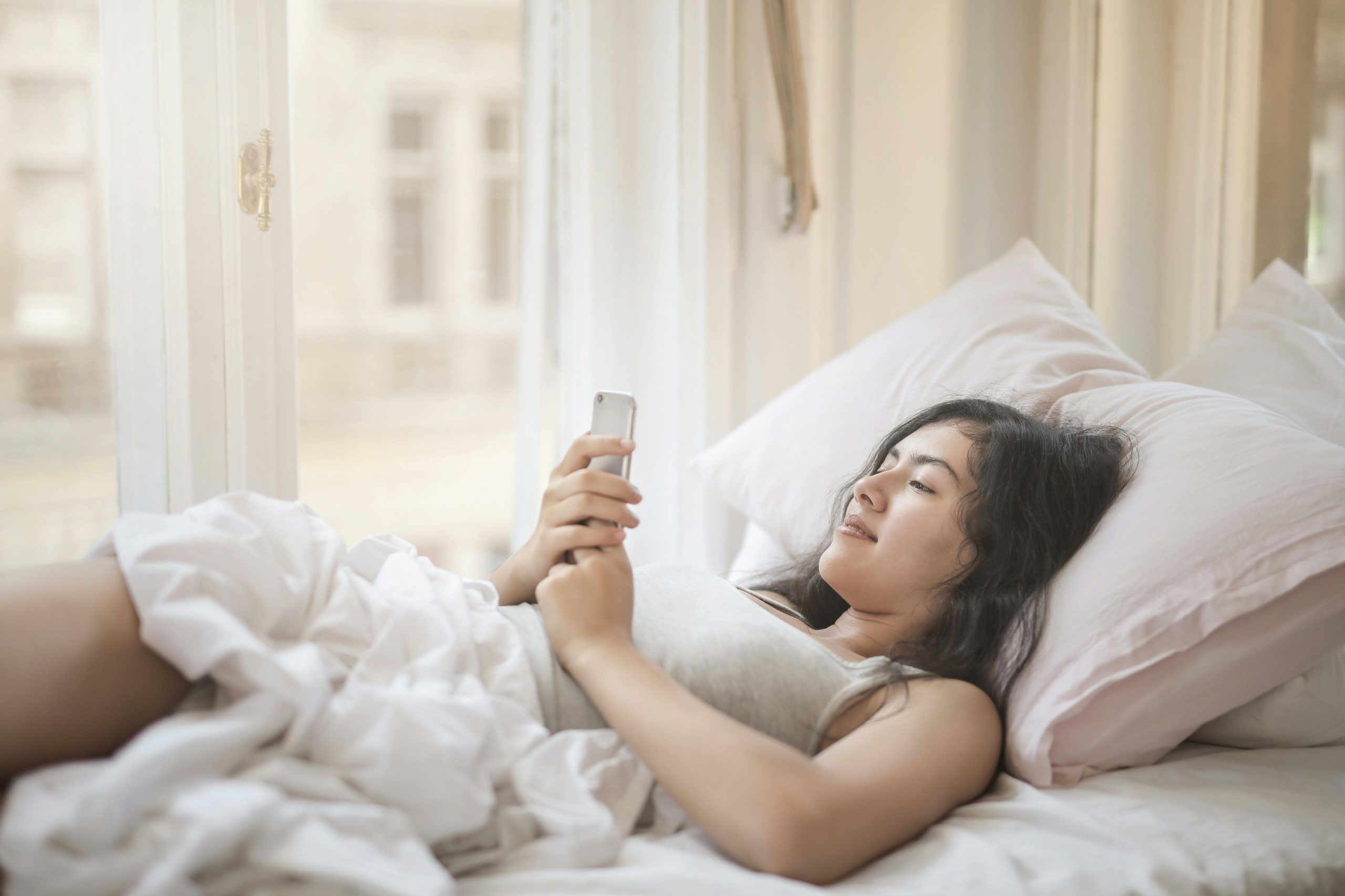 smiling woman relaxing in bed while using a smartphone